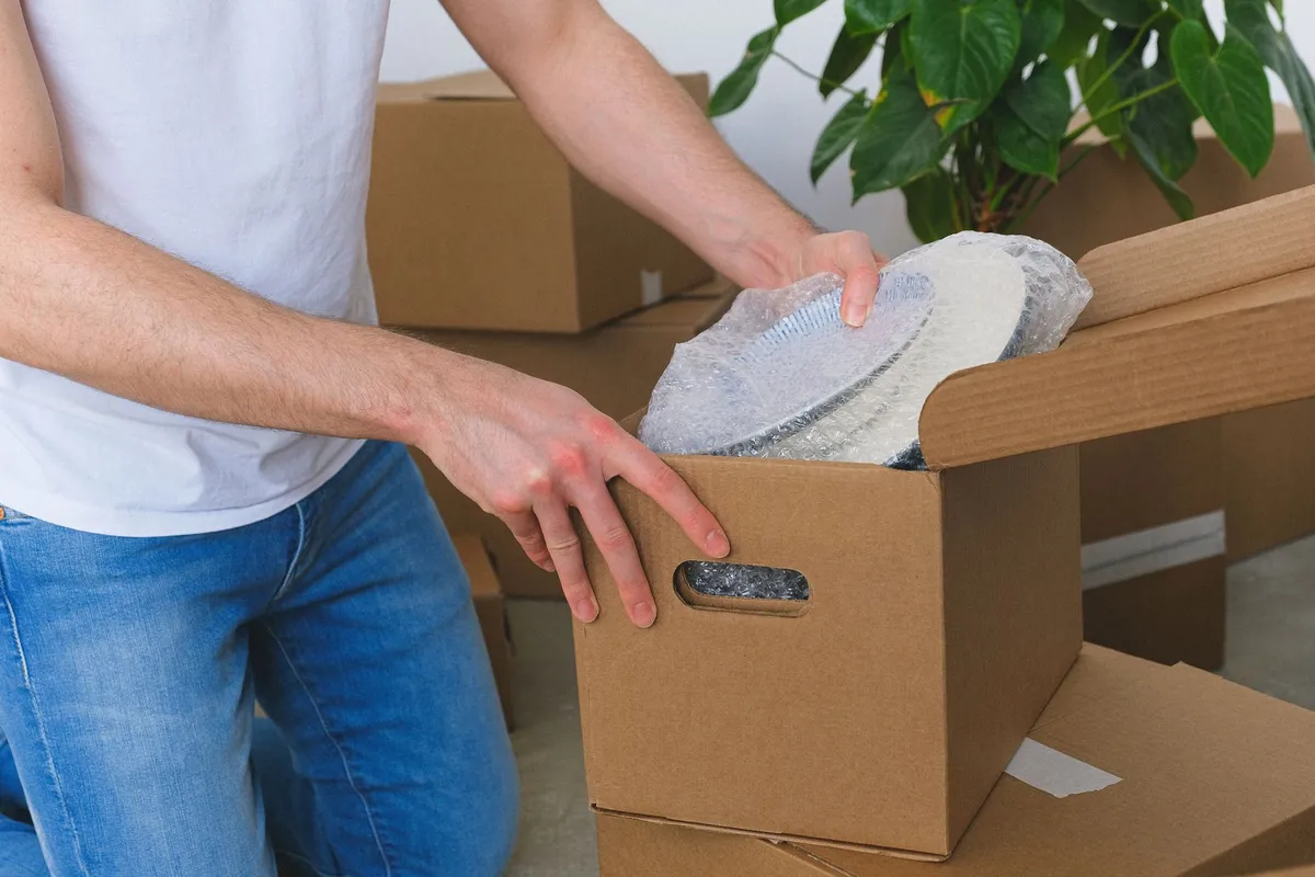 Mann verpackt einen in Luftpolsterfolie gewickelten Teller in einen unbedruckten Umzugskarton