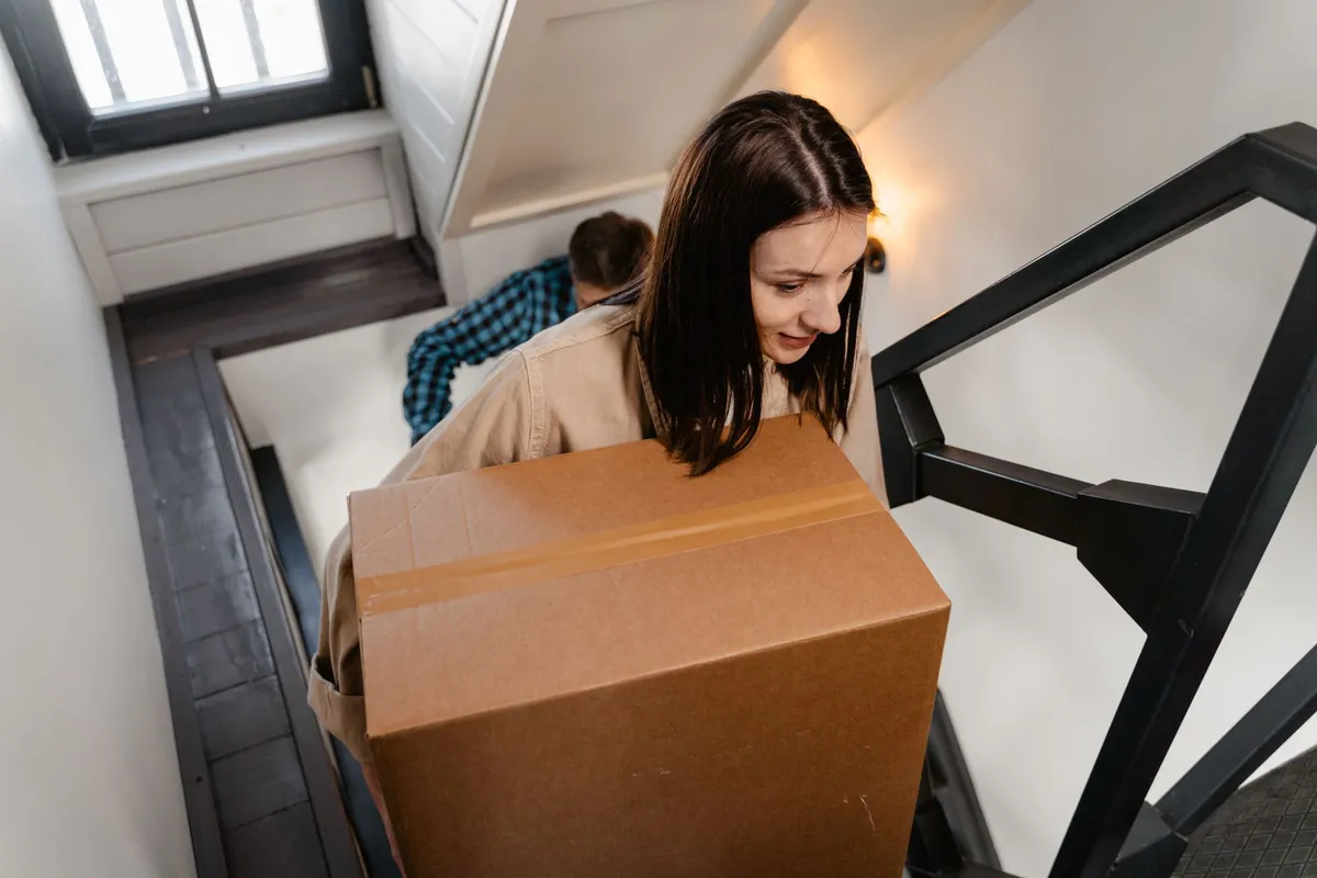 Helferin trägt einen großen Umzugskarton die Treppe hinauf, ein zweiter Helfer folgt