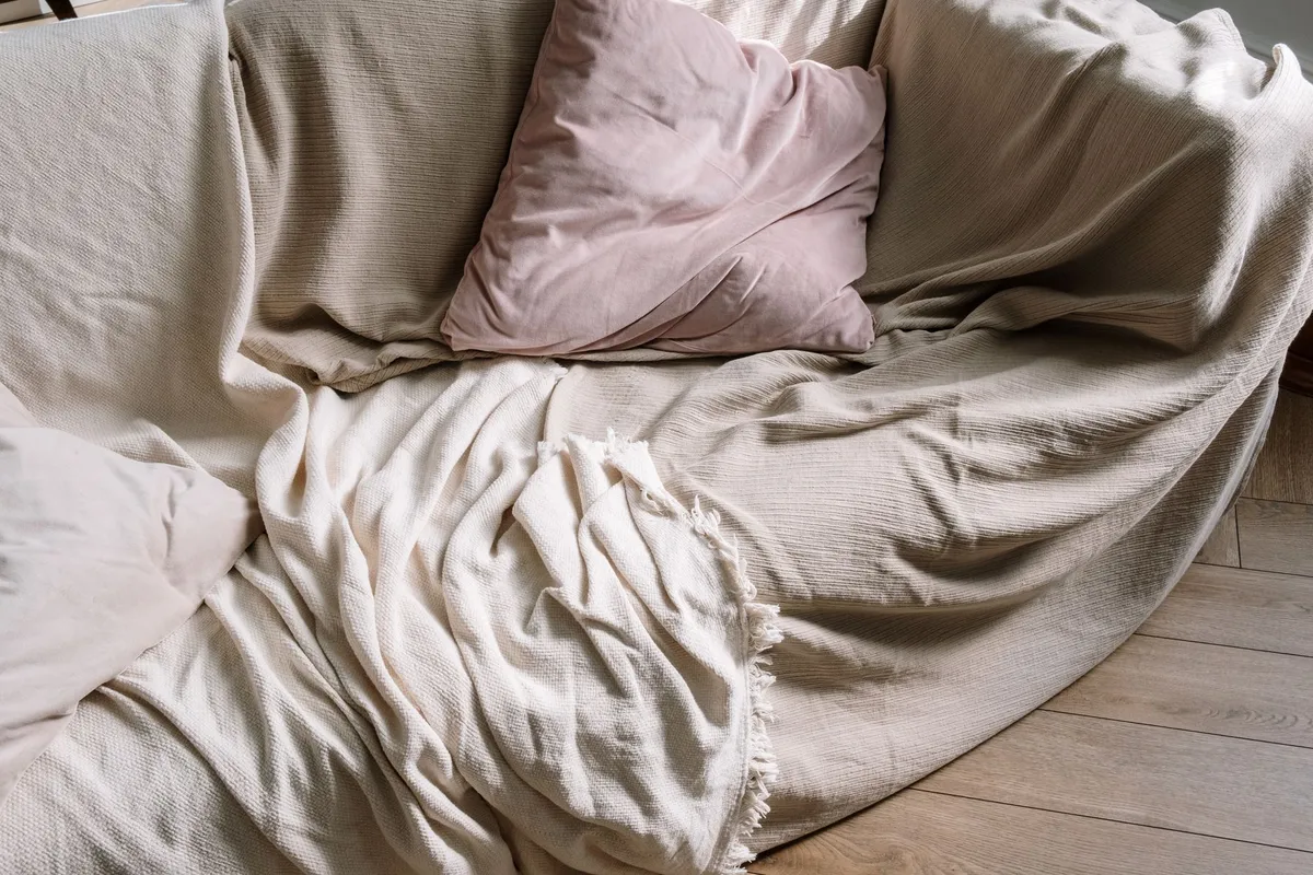 Sofa mit hellen Decken abgedeckt, davor ein Samtkissen und heller Dielenboden