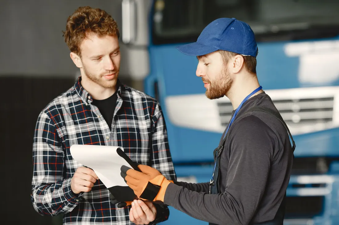 Fahrer mit Cap und Kollege besprechen vor einem blauen Lkw die Papiere auf dem Klemmbrett